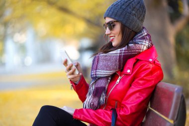 Bir bankta oturan ve sonbaharda cep telefonu kullanarak güzel genç kadın.