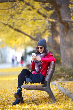 Bir bankta oturan ve sonbaharda cep telefonu kullanarak güzel genç kadın.