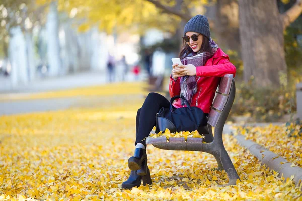 Hermosa joven sentada en un banco y usando su teléfono móvil en otoño . — Foto de Stock