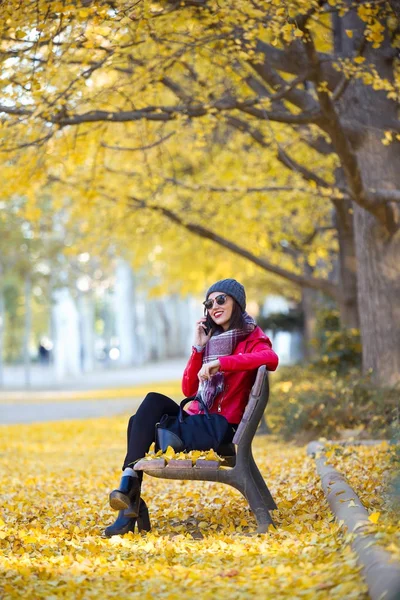 Vacker ung kvinna sitter i en bänk och använda sin mobiltelefon i höst. — Stockfoto