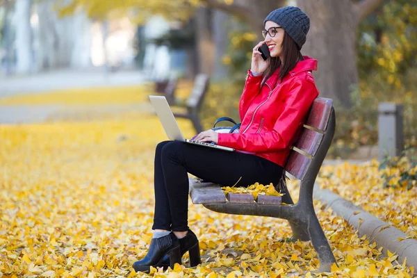 Hermosa mujer joven usando su teléfono móvil mientras trabaja con el ordenador portátil en otoño . —  Fotos de Stock