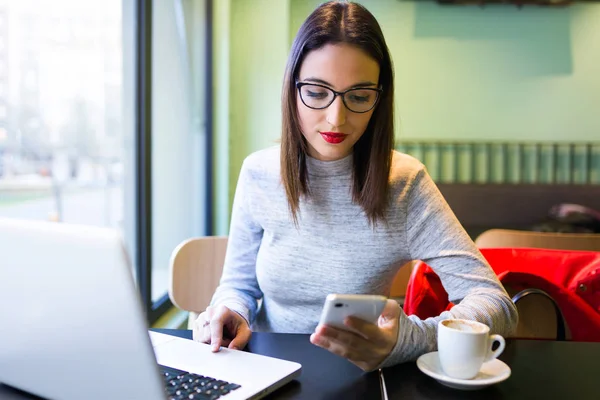 Schöne junge Frau mit Handy während der Arbeit mit ihrem Laptop im Café. — Stockfoto