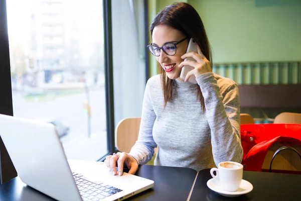 Bella giovane donna che utilizza il telefono cellulare mentre lavora con il suo computer portatile nella caffetteria . — Foto Stock