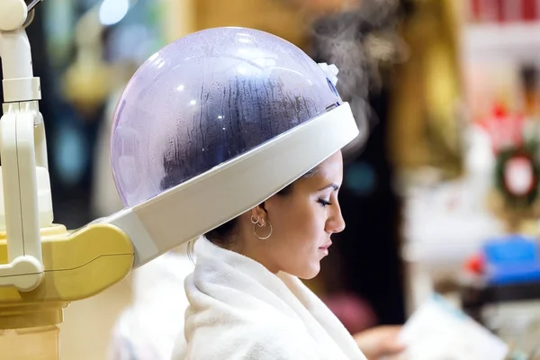 Hermosa mujer joven secando el cabello bajo la máquina en un salón de belleza . — Foto de Stock