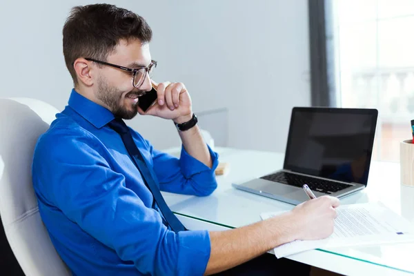 Joven guapo que trabaja con el ordenador portátil y el teléfono móvil en la oficina . — Foto de Stock