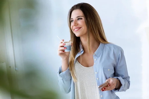 Mooie jonge vrouw drinken koffie tijdens het gebruik van haar mobiele telefoon thuis. — Stockfoto