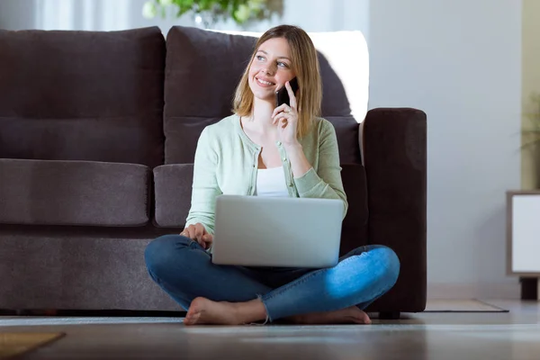 Vacker ung kvinna med hennes smartphone medan du arbetar med laptop hemma. — Stockfoto