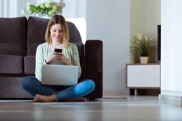 Schöne junge Frau, die ihr Smartphone benutzt, während sie zu Hause mit Laptop arbeitet. — Stockfoto