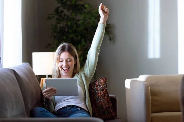 Schöne junge Frau feiert einen Erfolg, während sie ihr digitales Tablet zu Hause benutzt. — Stockfoto