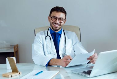 Erkek doktor ofiste onun hasta bilgileri yazılırken kameraya bakıyor.