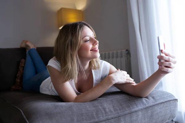 Hermosa joven feliz tomando una selfie con el teléfono móvil en casa . — Foto de Stock