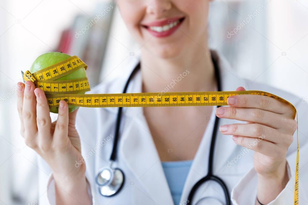 Young smiling female nutritionist holding a green apple wrapped in a tape measure in the consultation.