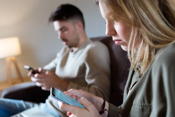 Hermosa pareja aburrida joven usando sus teléfonos inteligentes en casa . — Foto de Stock