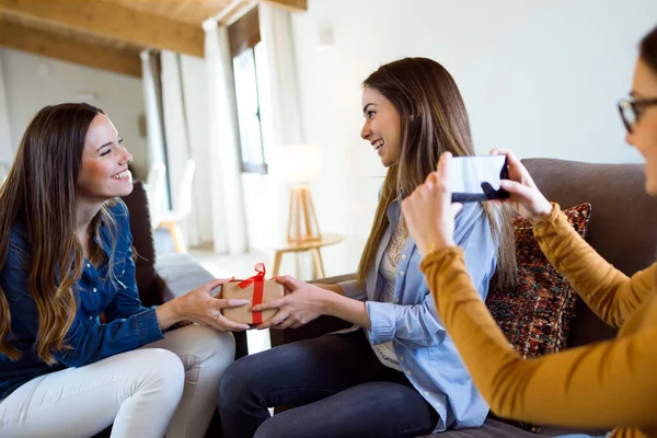 Two beautiful young women exchanging a gift while their friend takes a photo at home. — Stock Photo, Image