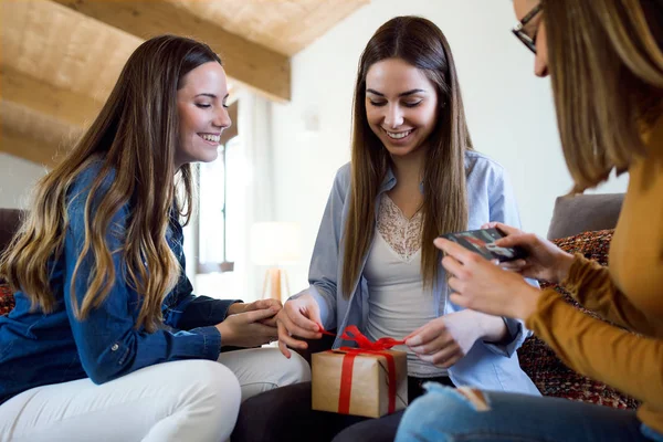 Dos hermosas mujeres jóvenes intercambiando un regalo mientras su amigo toma una foto en casa . — Foto de Stock