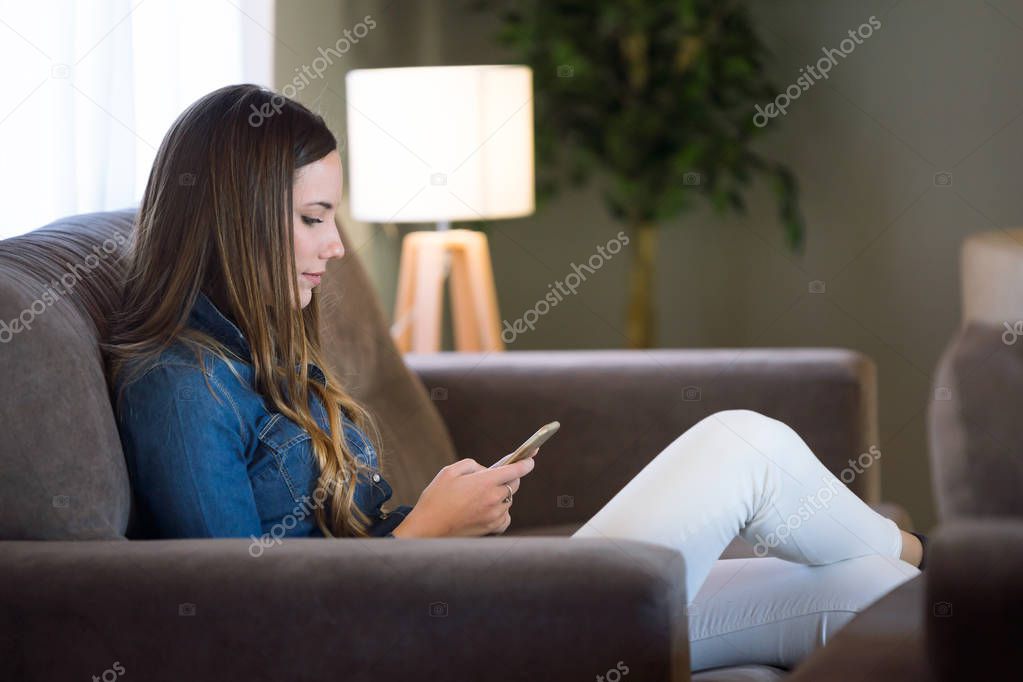 Beautiful young woman using her mobile phone and sitting on sofa at home.