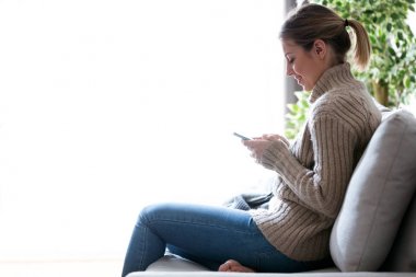 Evde kanepede otururken cep telefonu kullanan genç ve güzel kadın.