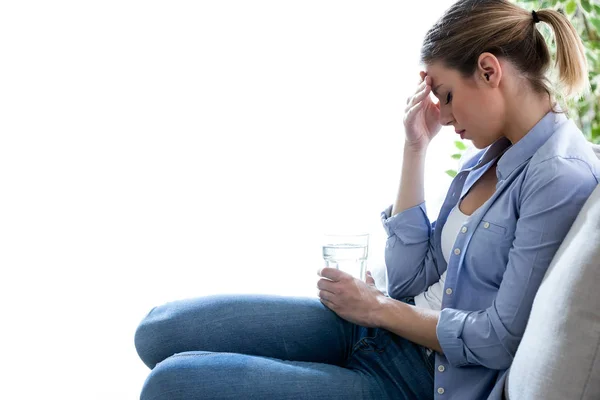 Niezdrowe młoda kobieta z bólem głowy, trzymając szklankę wody, siedząc na kanapie w domu. — Zdjęcie stockowe