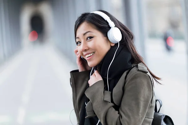 Sorrindo asiático jovem mulher ouvindo música e olhando para câmera na rua . — Fotografia de Stock