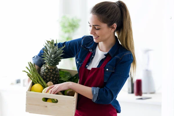 Mulher bonita segurando uma caixa com frutas e legumes na cozinha da casa . — Fotografia de Stock