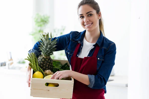 Mulher bonita segurando uma caixa com frutas e legumes na cozinha da casa . — Fotografia de Stock