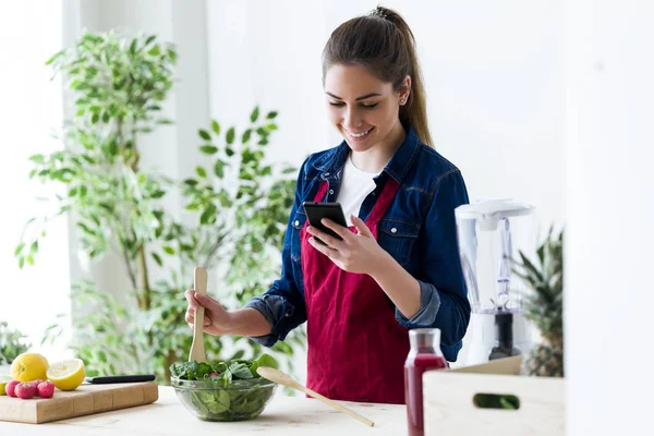 Mulher bonita usando seu telefone celular enquanto prepara salada na cozinha em casa . — Fotografia de Stock