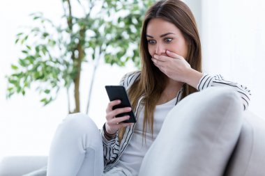 Evde kanepenin üzerinde cep telefonu kullanan sürpriz genç kadın.