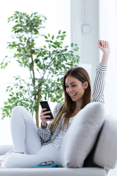 Bella giovane donna che celebra un successo mentre utilizza il suo telefono cellulare sul divano di casa . — Foto Stock