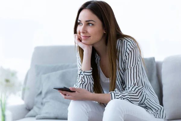 Hübsche junge Frau schaut seitwärts, während sie ihr Handy auf dem heimischen Sofa benutzt. — Stockfoto