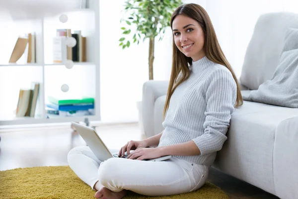 Bella giovane donna che guarda la fotocamera mentre lavora con il computer portatile a casa . — Foto Stock