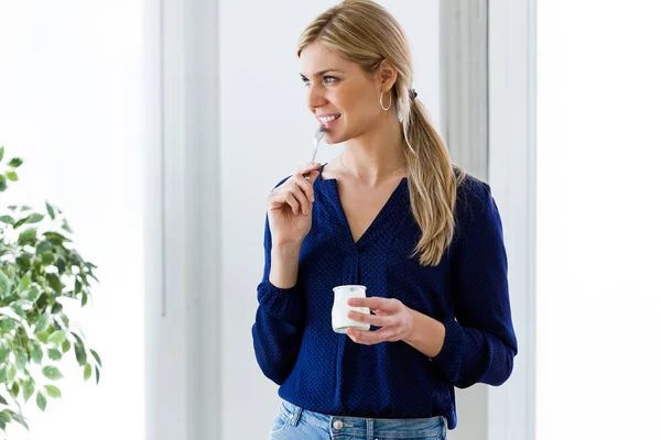 Hermosa mujer joven mirando hacia los lados mientras come yogur en casa . — Foto de Stock