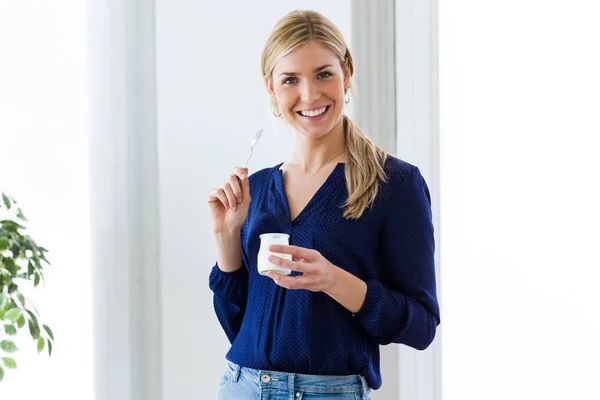 Mooie jonge vrouw camera kijken tijdens het eten van yoghurt thuis. — Stockfoto