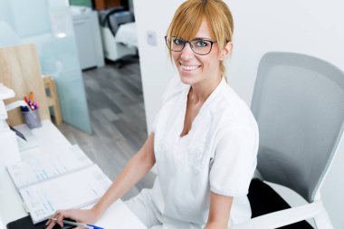 Kameraya bakarak ve klinik resepsiyonda gülümseyen genç ve güzel resepsiyonist.