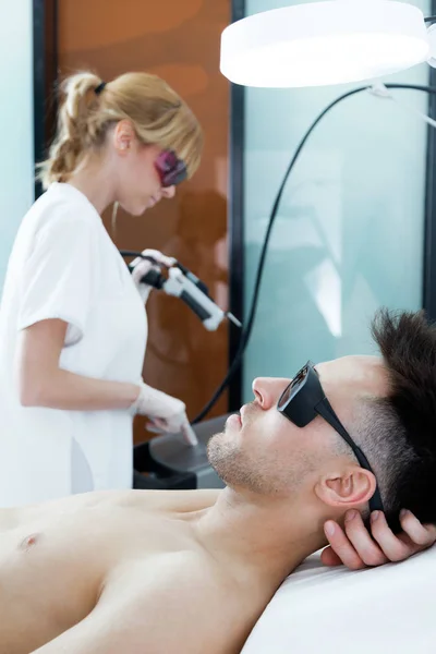 Handsome young man lying while the beautician adjusts the laser hair removal machine in beauty salon. — Stock Photo, Image