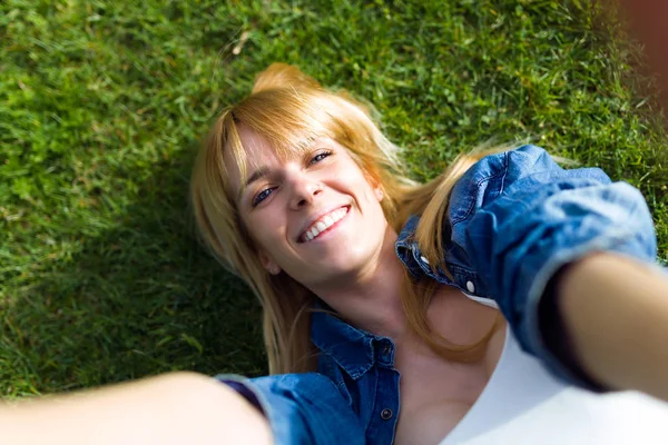 Mujer joven y bonita tomando una selfie mientras está tumbada en la hierba en el parque . —  Fotos de Stock