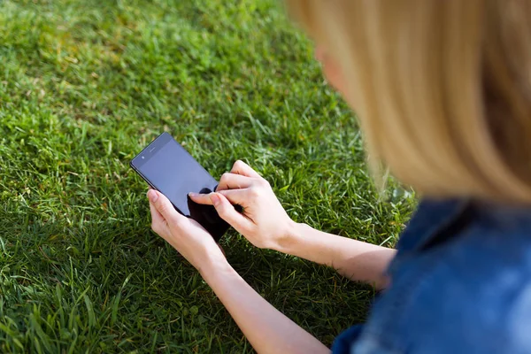 Hübsche junge Frau benutzt ihr Handy, während sie im Park auf Gras liegt. — Stockfoto