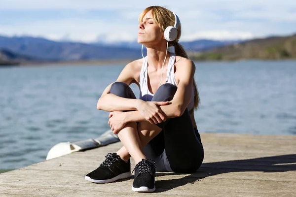 Fitte und sportliche junge Frau entspannt sich nach der Arbeit am See und hört Musik. — Stockfoto