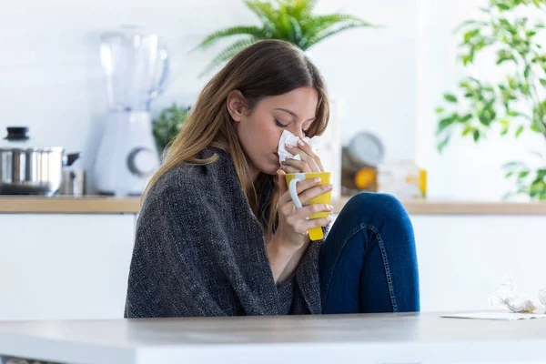 Doença jovem mulher espirrando em um tecido enquanto bebe bebida quente na cozinha em casa . — Fotografia de Stock