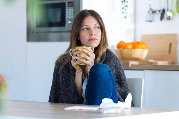 Sick and cold young woman drinking hot beverage while sitting in the kitchen at home. — Stock Photo, Image
