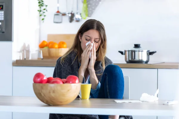 Krankheit junge Frau niest in ein Gewebe, während sie zu Hause in der Küche sitzt. — Stockfoto