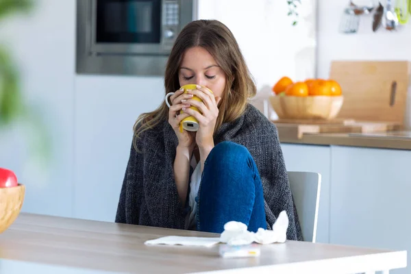Mujer joven enferma y fría bebiendo bebida caliente mientras está sentada en la cocina en casa . — Foto de Stock