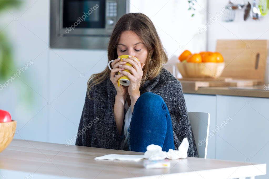 Sick and cold young woman drinking hot beverage while sitting in the kitchen at home.