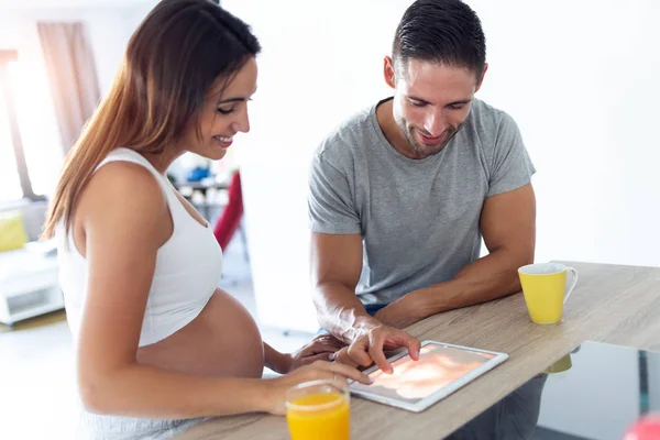 Hübsches junges schwangeres Paar betrachtet daheim in der Küche den Ultraschall ihres Babys auf dem digitalen Tablet. — Stockfoto