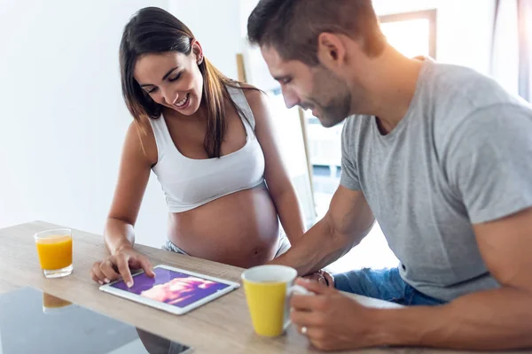 Muito jovem casal grávida olhando para o ultra-som de seu bebê no tablet digital na cozinha em casa . — Fotografia de Stock