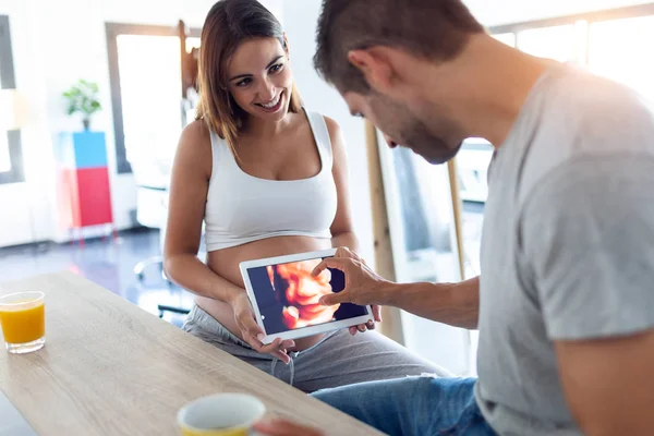Muito jovem grávida segurando o tablet digital com o ultra-som de seu bebê, enquanto o marido olhando isso em casa . — Fotografia de Stock