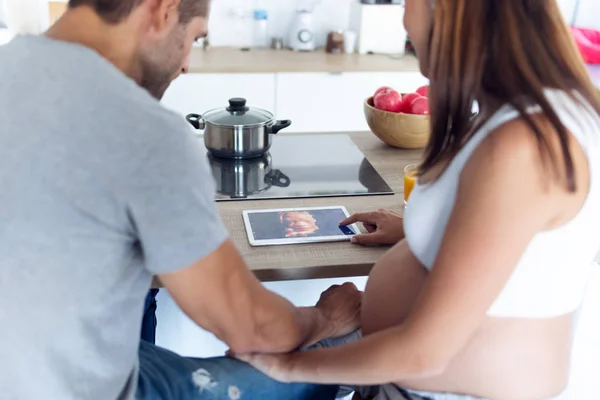 Hübsches junges schwangeres Paar betrachtet daheim in der Küche den Ultraschall ihres Babys auf dem digitalen Tablet. — Stockfoto