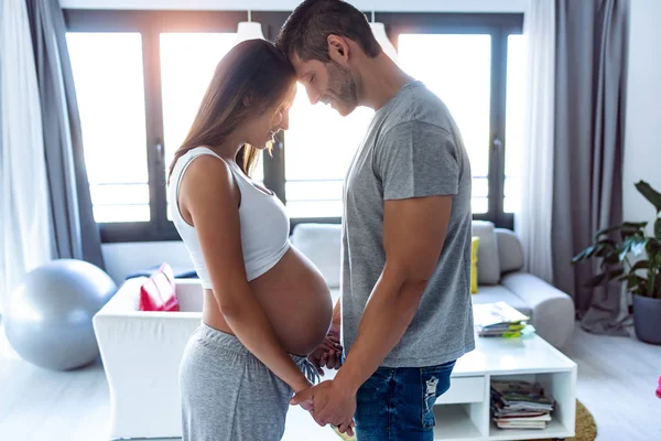 Hübsches junges schwangeres Paar blickt auf ihren Bauch, während es sich im heimischen Wohnzimmer an den Händen hält. — Stockfoto