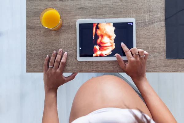 Mujer embarazada mirando y tocando la tableta digital con el ultrasonido de su bebé en casa . —  Fotos de Stock