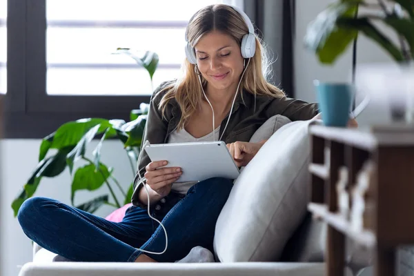 Hübsche junge Frau hört Musik mit Kopfhörern und ihrem digitalen Tablet, während sie zu Hause auf dem Sofa sitzt — Stockfoto