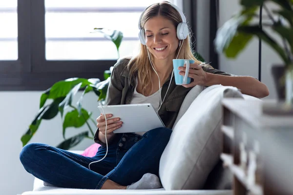 Mujer bastante joven escuchando música con su tableta digital mientras bebe una taza de café en el sofá en casa . — Foto de Stock